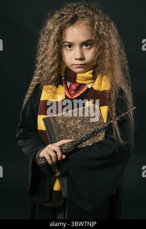 Portrait of a cute girl sorcière avec de magnifiques longs cheveux bruns vêtu d'un manteau foncé, jaune et bordeaux foulard rayé. Elle tient une baguette magique Banque D'Images