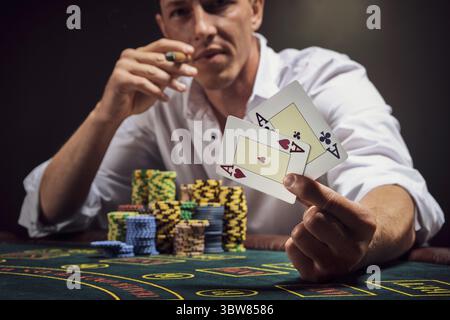 Athlétique homme dans une chemise blanche joue au poker assis à la table au casino en fumée, contre un projecteur blanc. Il fume un cigare en montrant Banque D'Images