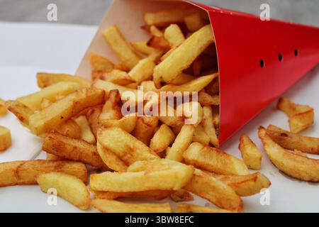 Frites belges dorées dans un cône de papier rouge, capturées à l'extérieur. Une collation de rue européenne classique à la lumière naturelle. Banque D'Images