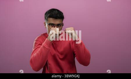 Jeune homme posant avec confiance en chemise rouge sur un fond rose vif, portant des lunettes et démontrant une position de boxe ludique. Banque D'Images
