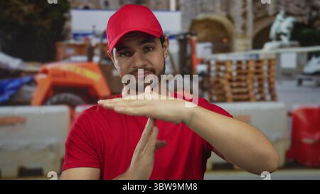 L'homme en chemise rouge fait signe de temporisation avec les deux mains au bâtiment en construction à l'extérieur à la lumière du jour ; pause. Banque D'Images