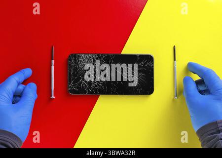 Les mains gantées d'un réparateur de quincaillerie à côté de tournevis et d'un téléphone cassé sur un fond jaune-rouge. Vue de dessus avec espace de copie Banque D'Images