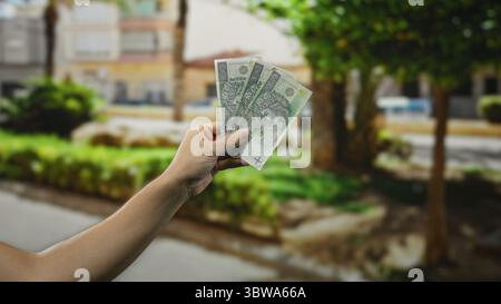 Homme tenant des billets de banque en zloty polonais sur une rue ensoleillée de la ville, présentant le change de devises et des concepts financiers à l'extérieur avec des arbres et des bâtiments en th Banque D'Images