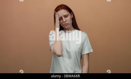 Femme en chemise blanche avec les cheveux rouges semble affligée sur fond brun, exprimant l'inconfort avec la main sur le front, cadre isolé. Banque D'Images