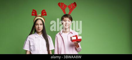 Couple d'adolescents caucasiens souriants dans la tenue de noël avec des cornes de cerf sur leurs têtes tenant une boîte cadeau et souriant joliment sur fond de studio vert. EMO Banque D'Images