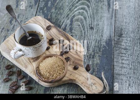 Café noir dans une vieille tasse de café avec une cuillère à café en argent, grains de café renversés et sucre de canne dans un bol en bois sur la vieille table en bois Banque D'Images