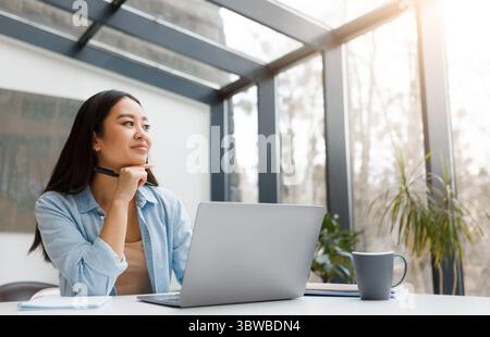 Femme asiatique réfléchie avec ordinateur portable dans l'espace de travail ensoleillé Banque D'Images