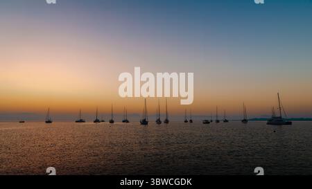 Un coucher de soleil serein sur une mer calme avec des silhouettes de voiliers à l'horizon. Banque D'Images