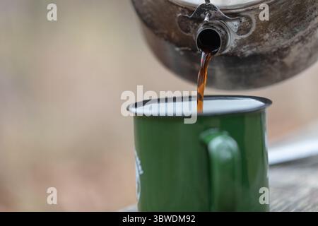 Gros plan du café frais versé de la bouilloire en métal dans la tasse de camping en émail vert, montée de vapeur, arrière-plan extérieur naturel flou. Banque D'Images