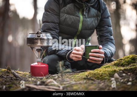 La personne en veste duvet est assise sur le sol de la forêt tenant la tasse de camping verte, bouilloire en métal sur la cuisinière à gaz au premier plan, fond boisé flou. Banque D'Images
