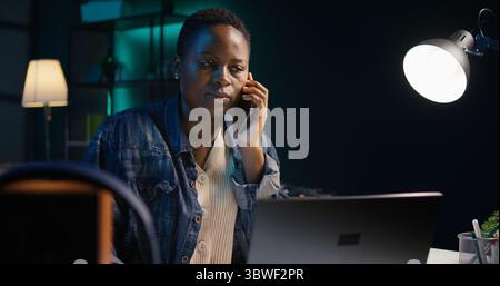 Femme recevant l'appel téléphonique d'un ami tout en travaillant de la maison sur ordinateur portable dans le bureau personnel. Freelance parlant avec mate en ligne à l'aide du téléphone portable tout en résolvant des tâches, caméra B. Banque D'Images