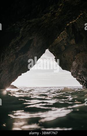 Une grande grotte rocheuse avec une petite ouverture au milieu. L'eau est calme et le ciel nuageux Banque D'Images