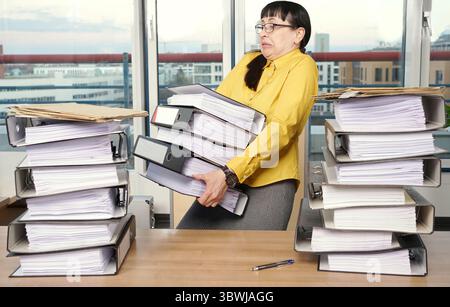 Secrétaire porte des dossiers lourds et trop nombreux, il y en a encore plus sur le bureau Banque D'Images