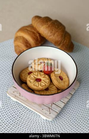 Gros plan d'un bol rose avec des biscuits sandwich à la confiture et une fraise fraîche sur le dessus. Deux croissants créent un fond chaleureux pour ce petit déjeuner sucré. Banque D'Images