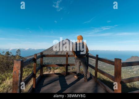 Un randonneur asiatique en veste jaune et sac à dos admire le volcan du mont Inerie depuis un point de vue en bois pittoresque à l'est de Nusa Tenggara, en Indonésie. Banque D'Images