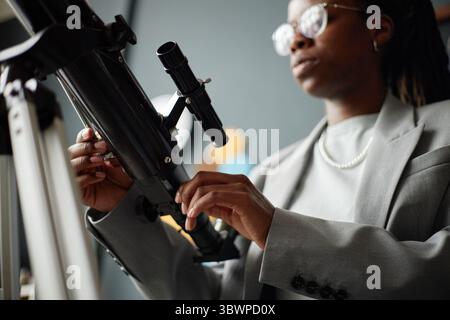 Jeune adulte femme noire ajustant télescope avec une expression focalisée, portant des lunettes et un costume, debout à l'intérieur, les mains manipulant soigneusement l'équipement, arrière-plan flou visible Banque D'Images