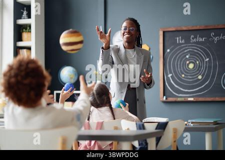 Enseignante noire engageant un groupe d'enfants dans une classe de sciences, jetant des planètes modèles tandis que les enfants atteignant pour les attraper, diagramme du système solaire visible sur le tableau noir en arrière-plan Banque D'Images