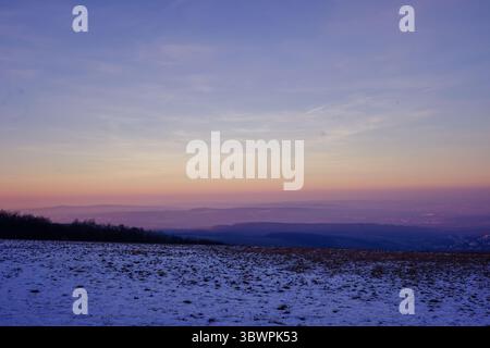 Paysage d'hiver avec champ enneigé et collines superposées au coucher du soleil, avec un ciel dégradé dans des couleurs pastel douces de l'orange au violet. Calme et atmosphe Banque D'Images