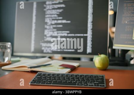 Gros plan montrant le clavier sans fil sur le bureau avec ordinateur portable, stylo, pomme verte, verre d'eau et moniteurs d'ordinateur affichant le code de programmation, configuration de l'espace de travail pour le développement de logiciels Banque D'Images
