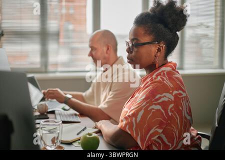 Jeune femme adulte noire portant des lunettes travaillant au bureau à côté d'un homme caucasien utilisant un ordinateur portable dans un cadre de bureau moderne, les deux axées sur les écrans d'ordinateur, la lumière naturelle du jour diffusant à travers les fenêtres Banque D'Images