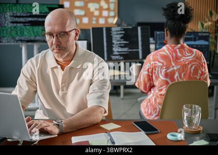 Homme caucasien d'âge moyen portant des lunettes travaillant sur ordinateur portable au bureau tandis que femme noire avec les cheveux bouclés assis à l'ordinateur en arrière-plan, codant dans l'espace de travail de bureau moderne Banque D'Images