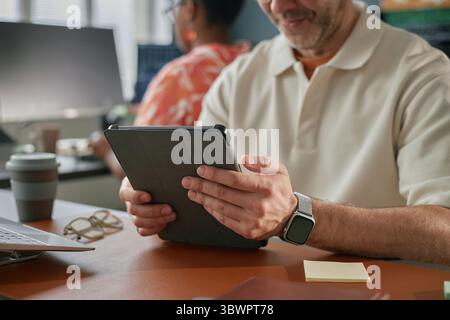 Homme caucasien d'âge moyen utilisant une tablette numérique au bureau dans un bureau moderne, assis près d'une femme noire travaillant à l'ordinateur, les deux axés sur les tâches, les appareils technologiques visibles sur l'espace de travail Banque D'Images