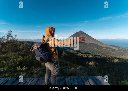 Un randonneur asiatique en veste jaune et sac à dos admire le volcan du mont Inerie depuis un point de vue en bois pittoresque à l'est de Nusa Tenggara, en Indonésie. Banque D'Images