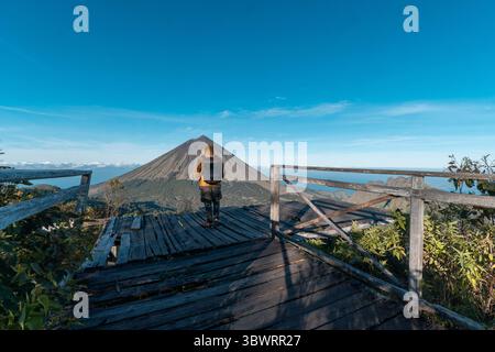 Un randonneur asiatique en veste jaune et sac à dos admire le volcan du mont Inerie depuis un point de vue en bois pittoresque à l'est de Nusa Tenggara, en Indonésie. Banque D'Images