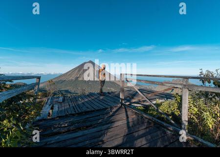 Un randonneur asiatique en veste jaune et sac à dos admire le volcan du mont Inerie depuis un point de vue en bois pittoresque à l'est de Nusa Tenggara, en Indonésie. Banque D'Images