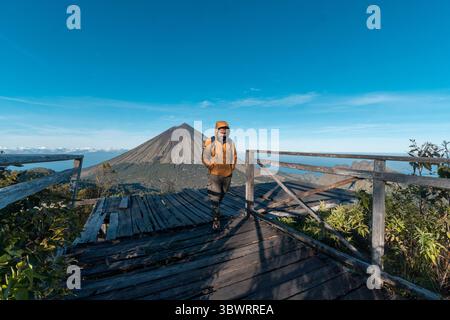 Un randonneur asiatique en veste jaune et sac à dos admire le volcan du mont Inerie depuis un point de vue en bois pittoresque à l'est de Nusa Tenggara, en Indonésie. Banque D'Images