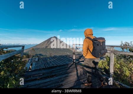 Un randonneur asiatique en veste jaune et sac à dos admire le volcan du mont Inerie depuis un point de vue en bois pittoresque à l'est de Nusa Tenggara, en Indonésie. Banque D'Images