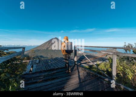 Un randonneur asiatique en veste jaune et sac à dos admire le volcan du mont Inerie depuis un point de vue en bois pittoresque à l'est de Nusa Tenggara, en Indonésie. Banque D'Images