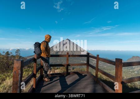 Un randonneur asiatique en veste jaune et sac à dos admire le volcan du mont Inerie depuis un point de vue en bois pittoresque à l'est de Nusa Tenggara, en Indonésie. Banque D'Images