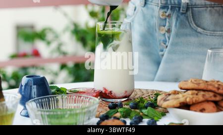 Femme versant le matcha fouetté dans un verre avec du lait, de la glace et de la confiture de baies. Préparation de latte matcha glacé en couches éclatante avec des ingrédients naturels frais Banque D'Images