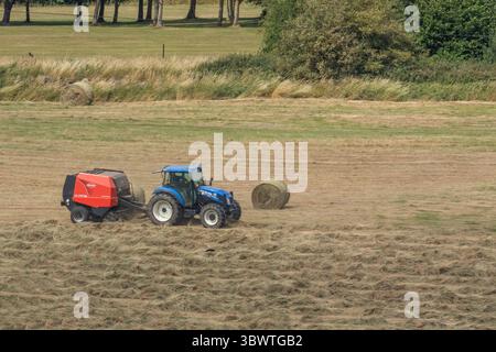 Tracteur bleu tirant une machine de mise en balles d'ensilage (presse à foin) dans le Yorkshire. L'herbe coupée est ramassée et transformée en balles prêtes à être entreposée. Banque D'Images