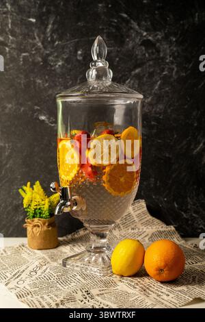 Élégant distributeur en verre d'eau infusée de fruits avec citron, orange et fraise. Set sur le journal avec des agrumes frais et des fleurs Solidago jaunes. Banque D'Images