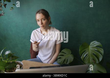 Jolie femme dans des lunettes et t-shirt blanc travaille sur ordinateur portable tout en étant assis dans une chaise à table à la maison. Fond vert foncé, à côté de lui est fleur en pot Banque D'Images