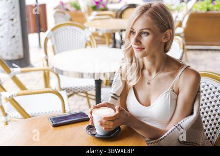 Jeune femme tout en se relaxant dans un café à table dans la rue, femme caucasienne heureuse est enveloppée dans un plaid chaleureux et confortable, quand elle boit du café dans un café en elle Banque D'Images