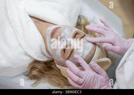 Masque de gommage facial, spa de beauté, soins de la peau. Femme obtenant le traitement du visage par esthéticienne au salon SPA, vue de côté, gros plan. Traitement anti-âge. Cosme Banque D'Images