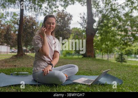 Femme assise à jambes croisées à l'extérieur parlant smartphone dispositif de maintien à l'oreille chemise pâle leggings gris ordinateur portable ouvert fond vert herbe Banque D'Images