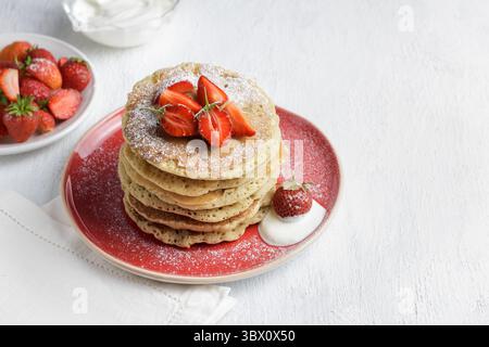 Pile de crêpes maison avec des fraises fraîches et du sucre en poudre sur l'assiette, servies avec du yaourt, gros plan sur fond blanc Banque D'Images