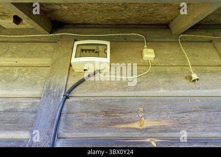 Une boîte électrique avec des fils et une prise montée sur un mur en bois altéré sous un simple plafond en bois, montrant l'utilité rustique. Banque D'Images