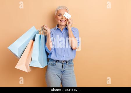 Femme âgée souriante profitant d'une journée de shopping tout en tenant des sacs colorés et en affichant une carte de crédit sur un fond beige Banque D'Images