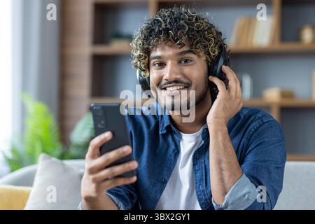 Un jeune homme souriant aime la musique sur son téléphone avec des écouteurs dans un cadre intérieur détendu. Banque D'Images