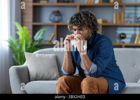 Un homme aux cheveux bouclés assis sur un canapé, se mouchant le nez, probablement éprouvant des allergies saisonnières ou un rhume. Banque D'Images