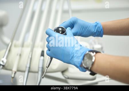 Gros plan d'un professionnel dentaire portant des gants en latex bleu tout en tenant et en inspectant une pièce à main dentaire dans une clinique moderne. Banque D'Images