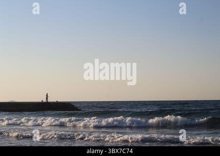 Une scène côtière tranquille au coucher du soleil met en scène un pêcheur solitaire debout sur une jetée rocheuse avec une canne à pêche. De douces vagues roulent sur le rivage sablonneux. Banque D'Images
