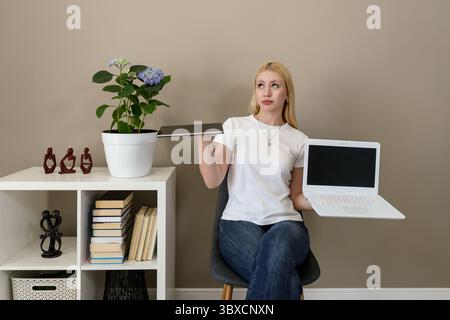 Femme blonde en tenue décontractée se trouve croisée avec une tablette dans une main et un ordinateur portable avec un écran blanc dans l'autre, regardant vers le haut réfléchi Banque D'Images