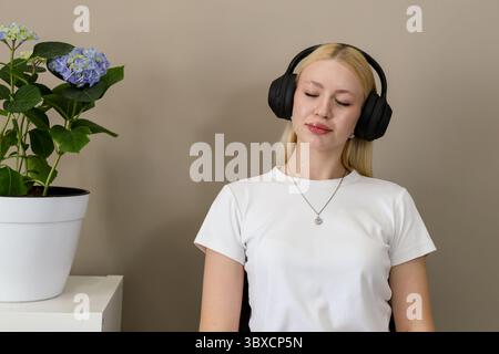 Jeune femme blonde dans un t-shirt blanc s'assoit paisiblement avec les yeux fermés, portant des écouteurs supra-auriculaires noirs et écoutant de la musique dans une pièce moderne et confortable Banque D'Images