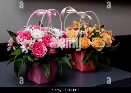 Beaux bouquets de fleurs mélangées avec des roses, des marguerites et de la verdure dans des paniers en papier pastel avec poignées en ruban, parfaits pour les fêtes ou les cadeaux Banque D'Images